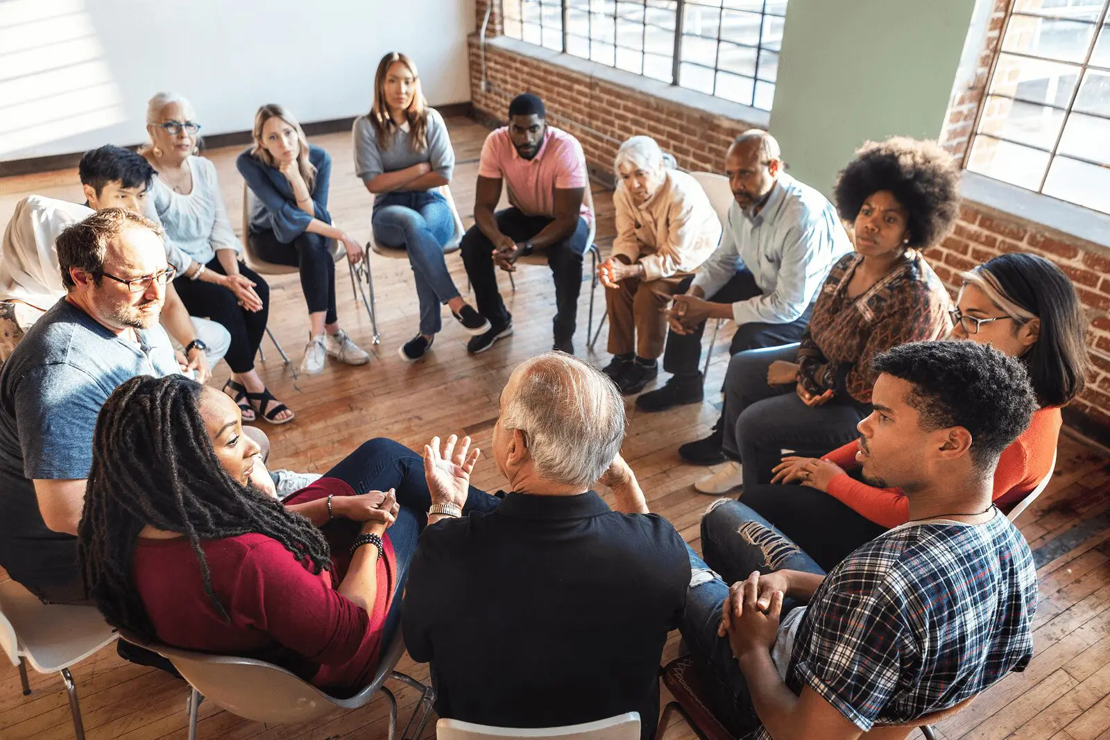 blog (1) A diverse group of people sit in a circle on chairs, engaged in conversation in a bright room with large windows and wooden floors.