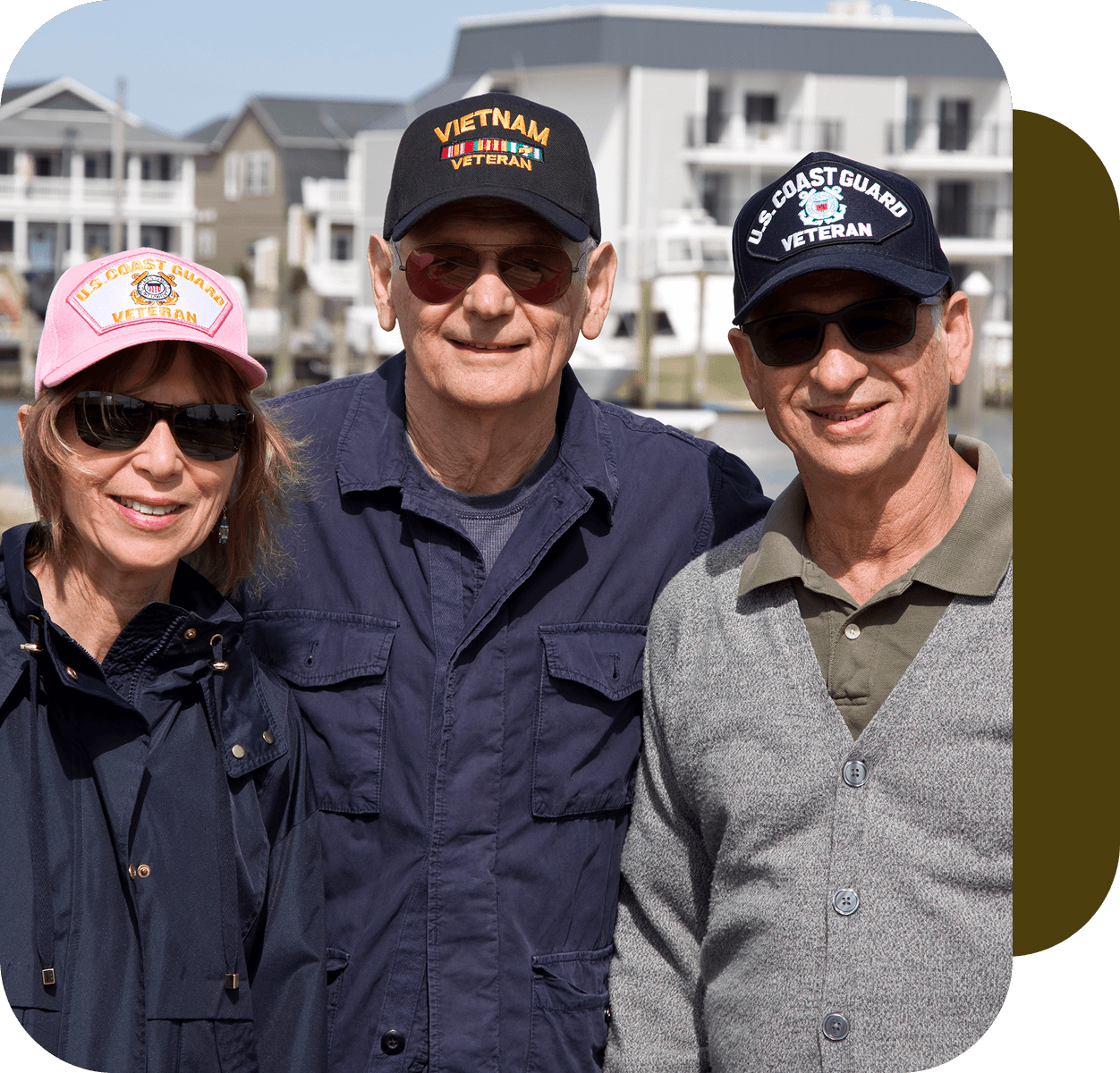 h2 (1) Three people outdoors wearing veteran caps, smiling; two men and one woman. Background features residential buildings.
