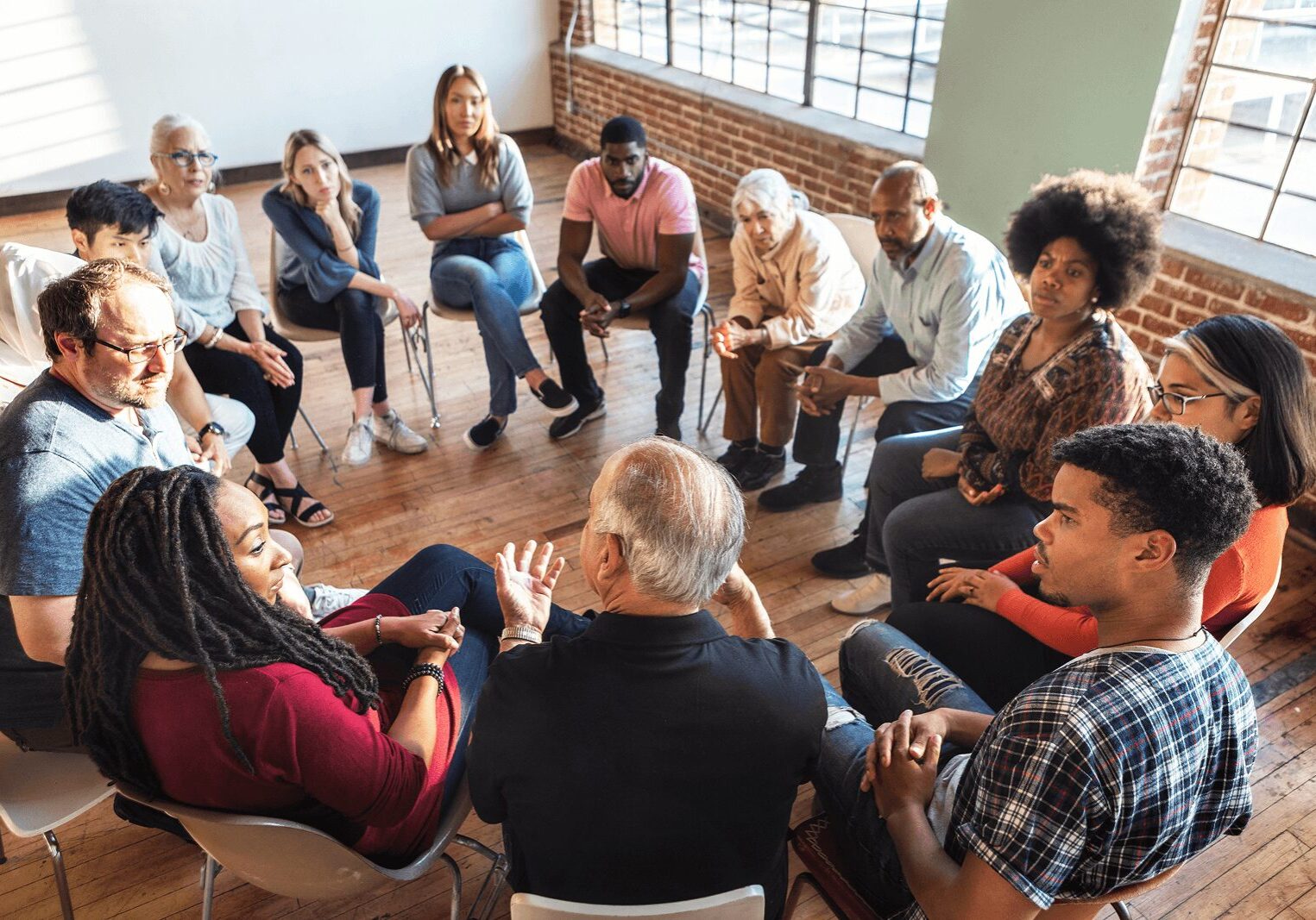 A diverse group of people sit in a circle on chairs, engaged in conversation in a bright room with large windows and wooden floors.